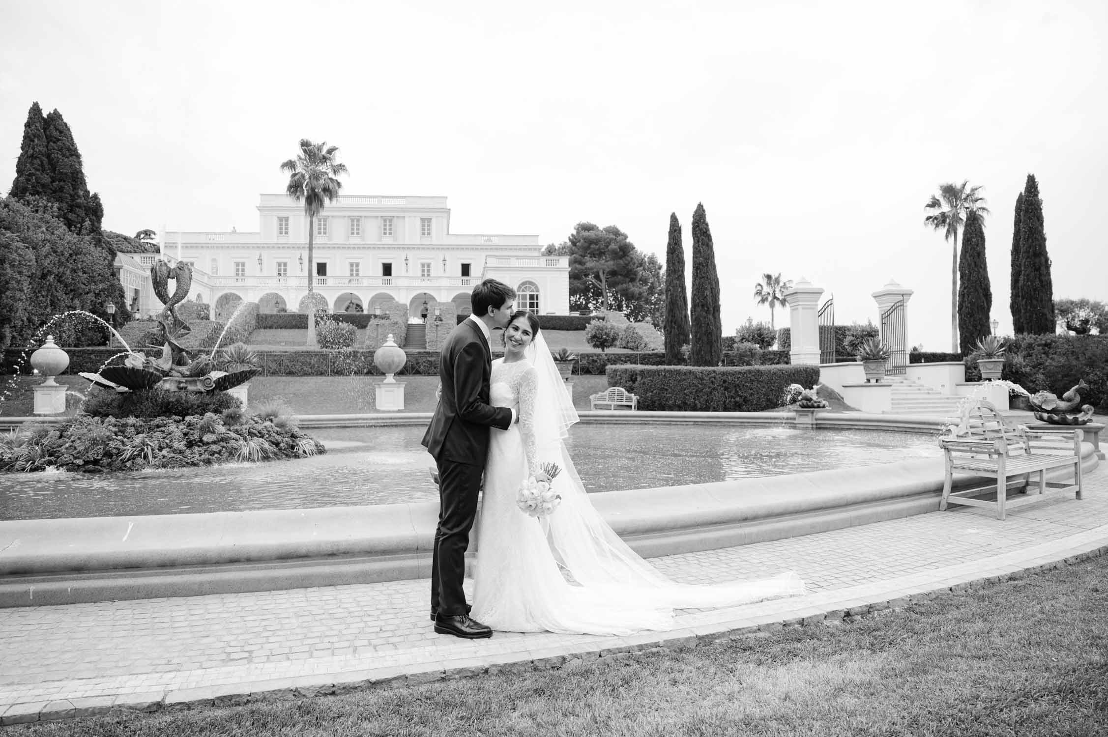 Bride and groom share a kiss in a formal estate garden with a grand villa, fountains, and trimmed hedges in the background (black and white).